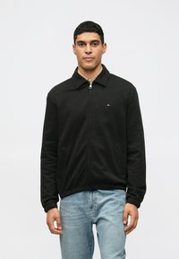 Crna jakna na zatvaranje s ovratnikom, koja ima bočne džepove, rebraste manžetne i mali logo na lijevoj strani prsiju. U kombinaciji s plavim trapericama.