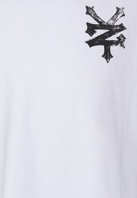 Hvid bomulds T-shirt med et sort grafikdesign på venstre bryst. Stoffet har en glat tekstur og en rund hals.