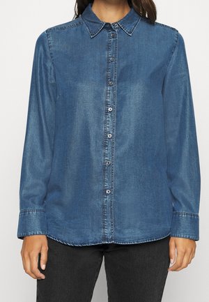 Vrouw die een blauwe denim blouse met knopen en lange mouwen draagt en een zwarte broek, staand tegen een effen lichte achtergrond.