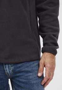 Tumši pelēks frotē jakas ar mīkstu tekstūru, sānu šuvju detaļu un elastīgām piedurknēm, apvienots ar ziliem džinsiem un roku, kas atpūšas uz augšstilba.