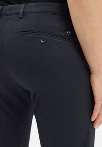 Zbliżenie na osobę w ciemnych, teksturowanych spodniach z zapięciem na guziki w tylnej kieszeni i ręką spoczywającą z boku.