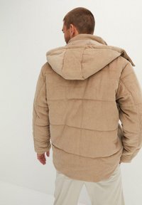 Giacca in corduroy beige con cappuccio, caratterizzata da un design trapuntato e una vestibilità rilassata. Texture a coste e dettagli di cucitura visibili sul retro.