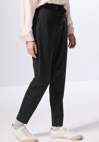 Svarte tapered bukser med snoreliv, med sidesømmer og en glat tekstur, parret med hvide sneakers og grå sokker.