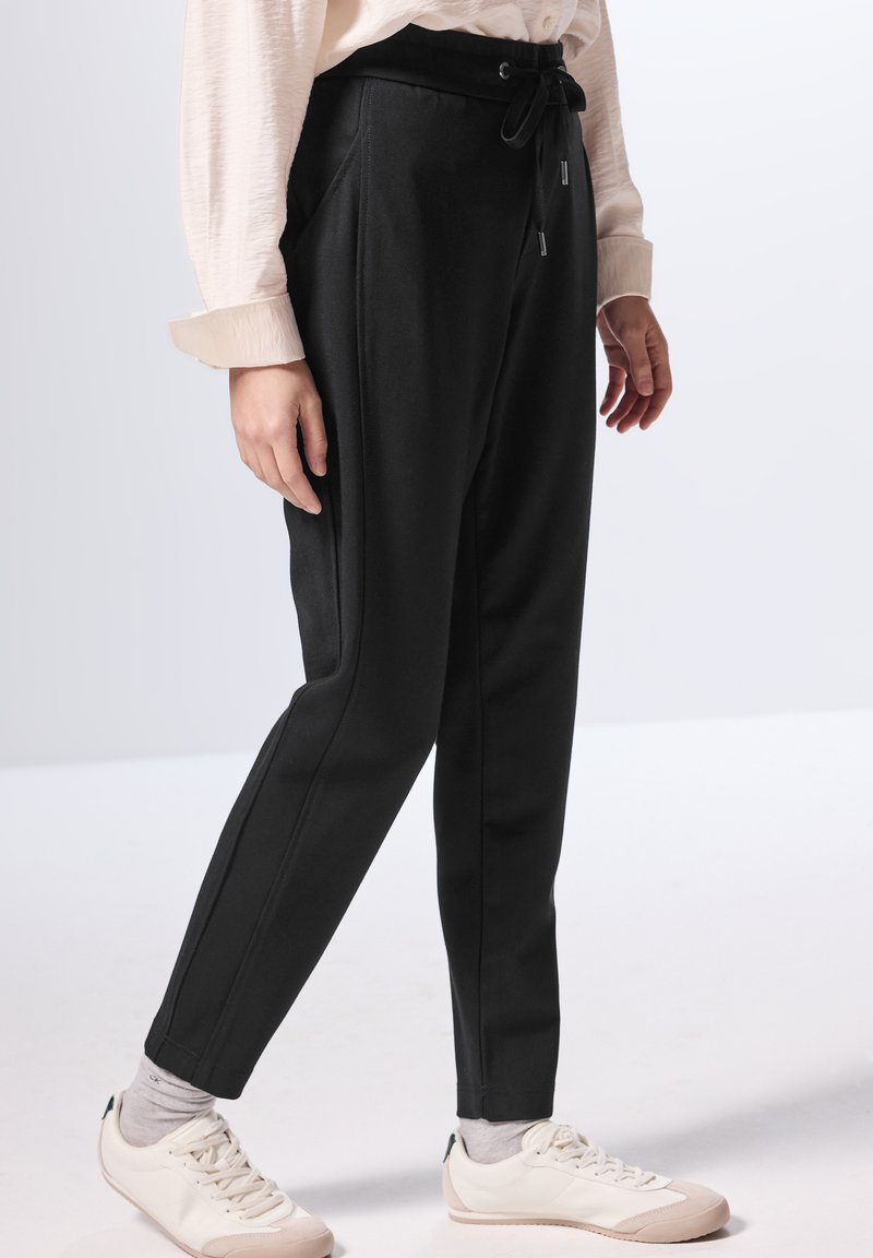 Svarte tapered bukser med snoreliv, med sidesømmer og en glat tekstur, parret med hvide sneakers og grå sokker.