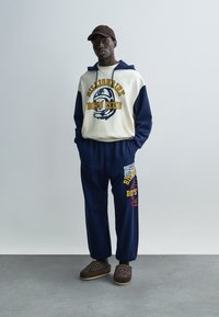 Μπλε ναυτικό και κρεμ φούτερ με κουκούλα με την επιγραφή "Billionaire Boys Club" σε κίτρινο. Μπλε ναυτικό παντελόνι φόρμας με πολύχρωμα γραφικά. Καφέ παντόφλες.