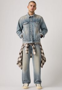 Ανοιχτό μπλε τζιν μπουφάν με σκούρες ραφές patchwork, κλείσιμο με κουμπιά και τσέπες στο στήθος, σε συνδυασμό με ανοιχτό μπλε τζιν παντελόνι και καρό πουκάμισο δεμένο στη μέση.