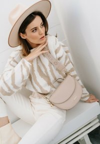 Sac en cuir beige en bandoulière avec un design incurvé et des accessoires en or, associé à un pull rayé crème et beige et un pantalon blanc.
