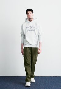 Siv hooded majica z logotipom 'New Balance Athletics', zeleni cargo hlače in bele športne copate. Model stoji ob preprosti steni.