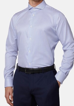 Hombre con una camisa blanca ajustada de vestir con finas rayas verticales azules, pantalones azul oscuro y un cinturón negro, una mano en el bolsillo.