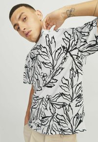 Kortærmet skjorte i hvidt stof med sort blad- og blomsterprint, der har en afslappet pasform og subtil tekstur, vist på en model.
