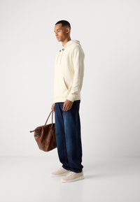 Mann in cremfarbener Kapuzenjacke, dunkelbblauen Jeans, beigen Sneakers, trägt eine große braune Ledertasche und steht vor einem schlichten weißen Hintergrund.