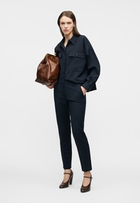 Γυναίκα με μπλε σκουρόχρωμο πουκάμισο και παντελόνι, κρατώντας μεγάλη καφέ δερμάτινη τσάντα, φορώντας καφέ ψηλοτάκουνα παπούτσια με λουράκια στον αστράγαλο, στέκεται μπροστά σε λευκό φόντο.