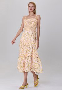 Robe maxi florale en rose clair avec des motifs de fleurs jaunes, corsage smocké et ourlet à volants superposés. Portée avec des sandales à talons jaunes.