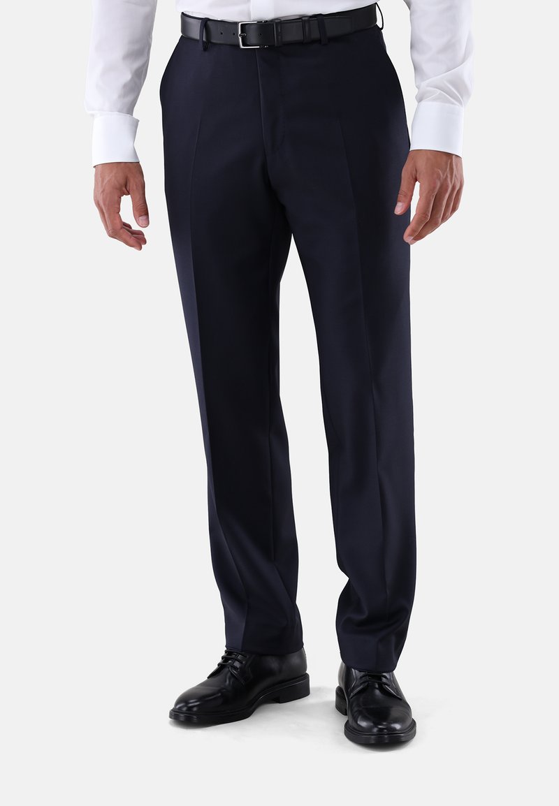 Mann trägt dunkelblaue Anzughose, schwarzen Gürtel, schwarze formelle Schuhe und weißes Hemd, steht vor einem einfarbigen Hintergrund.