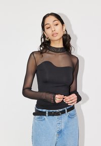 Top nero a maniche lunghe con un design in rete trasparente, colletto alto e corpetto aderente; abbinato a jeans blu a vita alta e una cintura nera.