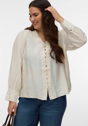 Uśmiechnięta kobieta poprawiająca włosy, ubrana w kremową bluzkę na guziki z bufkami na rękawach i niebieskie dżinsy, trzymająca brązową torebkę w lewej ręce.