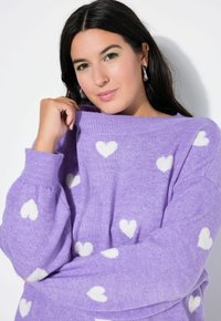 Lawendowy sweter z dzianiny z białymi wzorami serc, luźnym krojem, szerokim prążkowanym dekoltem i miękką fakturą. Długie rękawy z prążkowanymi mankietami.