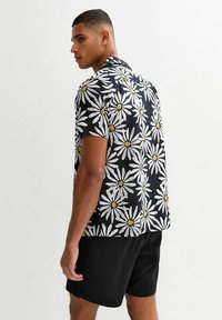 Chemise à manches courtes en tissu noir avec un motif de marguerites blanches et un centre jaune. Coupe régulière, texture subtile, avec un col.