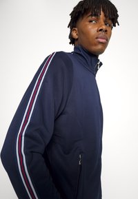 Giacca a zip blu navy con colletto alto, caratterizzata da strisce bianche, rosse e blu lungo le maniche e una tasca laterale con zip. Tessuto morbido.