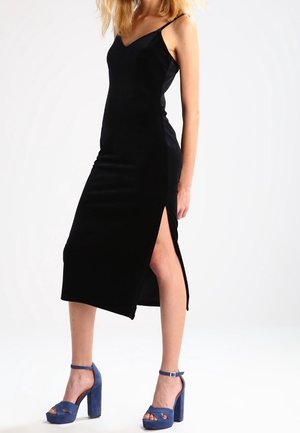 Schwarzes Samtkleid mit V-Ausschnitt und dünnen Trägern, mit einem seitlichen Schlitz bis zum Oberschenkel. Kombiniert mit blauen Plateau-Pumps mit Knöchelriemen.