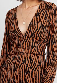 Donna che indossa un abito a V con maniche lunghe a strisce tigre marroni e nere, accompagnato da una collana d'argento con un ciondolo rotondo.