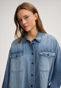 Light blue denim shirt featuring a button-up front, two chest pockets, and a classic collar. The fabric has a smooth texture with gradient shading.