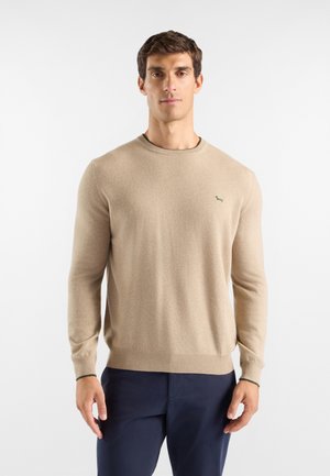 Uomo che indossa un maglione beige a maniche lunghe con collo tondo e pantaloni blu scuro, in piedi davanti a uno sfondo bianco uniforme.