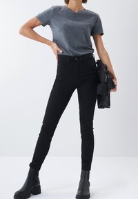 Jeans ajustados negros combinados con una camiseta de cuello redondo gris oscuro. Modelo llevando botas negras hasta el tobillo y sosteniendo un bolso negro. Textura suave.