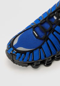 Modré sportovní boty s síťovinovým materiálem, černými akcenty, texturovanou podešví s bublinkovými prvky a aerodynamickým designem.