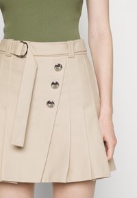 Jupe plissée beige avec trois boutons argentés décoratifs et une ceinture assortie, portée avec un haut côtelé vert olive.