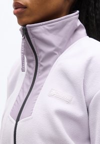 Κοντινό πλάνο ενός ατόμου που φοράει ένα ανοιχτό μωβ φλις μπουφάν Columbia με ψηλό γιακά και μαύρο φερμουάρ, μερικώς κλειστό.