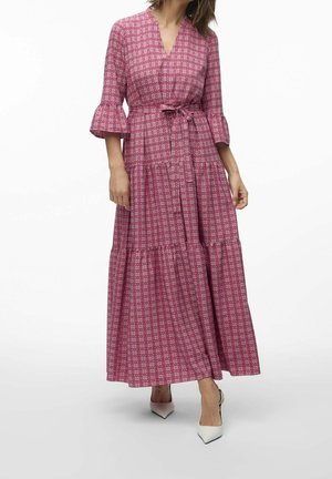 Femme portant une longue robe rose à motifs avec manches trois-quarts, taille ceinturée et talons blancs pointus sur un fond blanc uni.