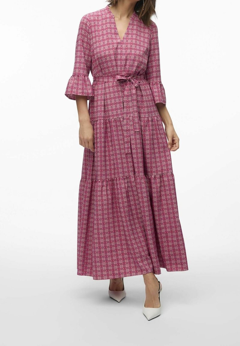 Femme portant une longue robe rose à motifs avec manches trois-quarts, taille ceinturée et talons blancs pointus sur un fond blanc uni.