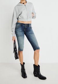 Grå kort hoodie med dragkedja och fickor, ihop med mörkblå denimbermuda-shorts och svarta ankelstövlar med silverdetaljer.