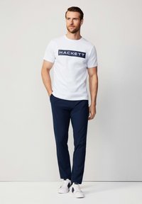 Camiseta blanca de manga corta con el logo "HACKETT", combinada con pantalones oscuros azules y zapatillas blancas con detalles en azul marino.