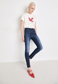 Cremfarbendes T-Shirt mit rotem Vogel-Graphic, kombiniert mit dunkelblauen Skinny Jeans. Das Model trägt rote spitze Flats mit zwei Riemen. Weißer Hintergrund.