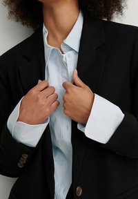Blazer nero con bottoni dorati indossato sopra una camicia azzurra chiaro. La camicia presenta un colletto classico e una chiusura a bottoni, con maniche arrotolate.
