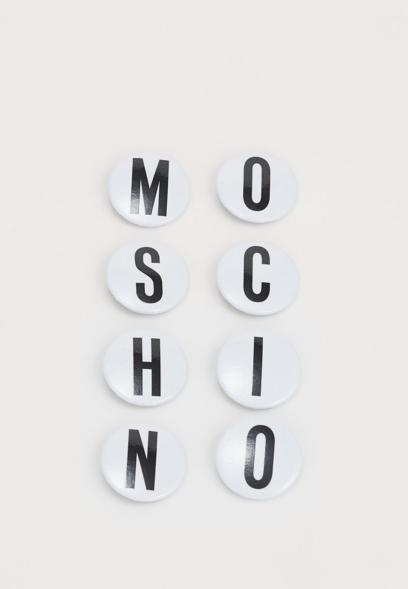 Astotne astoņu apaļu pogu komplekts ar melniem lielajiem burtiem uz baltas spīdīgas fona, kas veido vārdu "MOSCHINO" divās rindās.