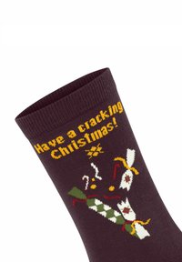 Dunkelbraune Socke mit dem gestrickten Text "Frohe Weihnachten!" und festlichen Designs, darunter Knallbonbons, musikalische Noten und bunte Akzente.