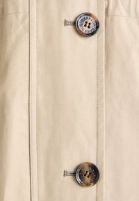 Μπεζ υφασμάτινο πουκάμισο με κουμπιά, που διαθέτει δύο μεγάλα, στρογγυλά κουμπιά με λογότυπο, επίπεδη ραφή και διακριτική γυαλάδα.