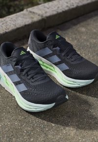 Μαύρα αθλητικά παπούτσια Adidas με ανοιχτό μπλε ρίγες και πράσινες μαλακές σόλες τοποθετημένα σε υφή εξωτερικής επιφάνειας κοντά σε μια πέτρινη άκρη.
