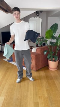 Model drži svetlo zeleno in črno prilegajočo srajco. Scena vključuje leseno tla, kavč z vertikalnimi črtami in lončnice.