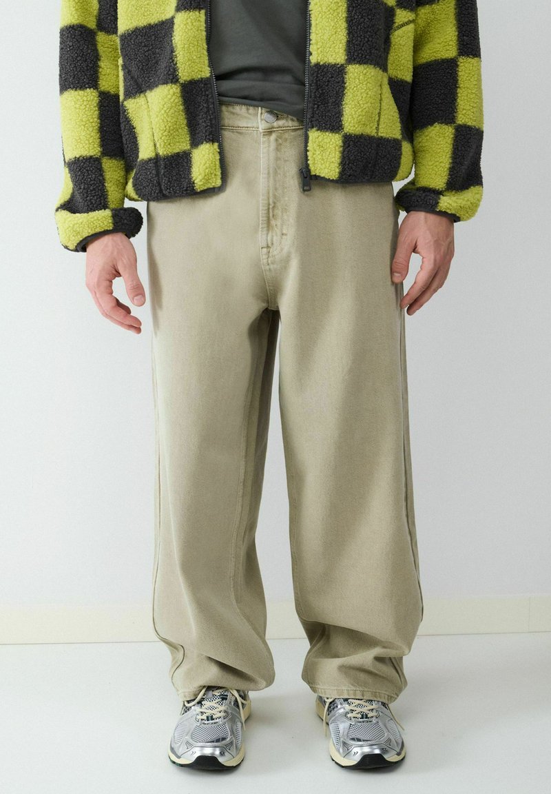 Beige wijde broek gecombineerd met een zwart en limoen groen geruite fleece jasje en zilveren sportsneakers. Zachte textuur en relaxte pasvorm.