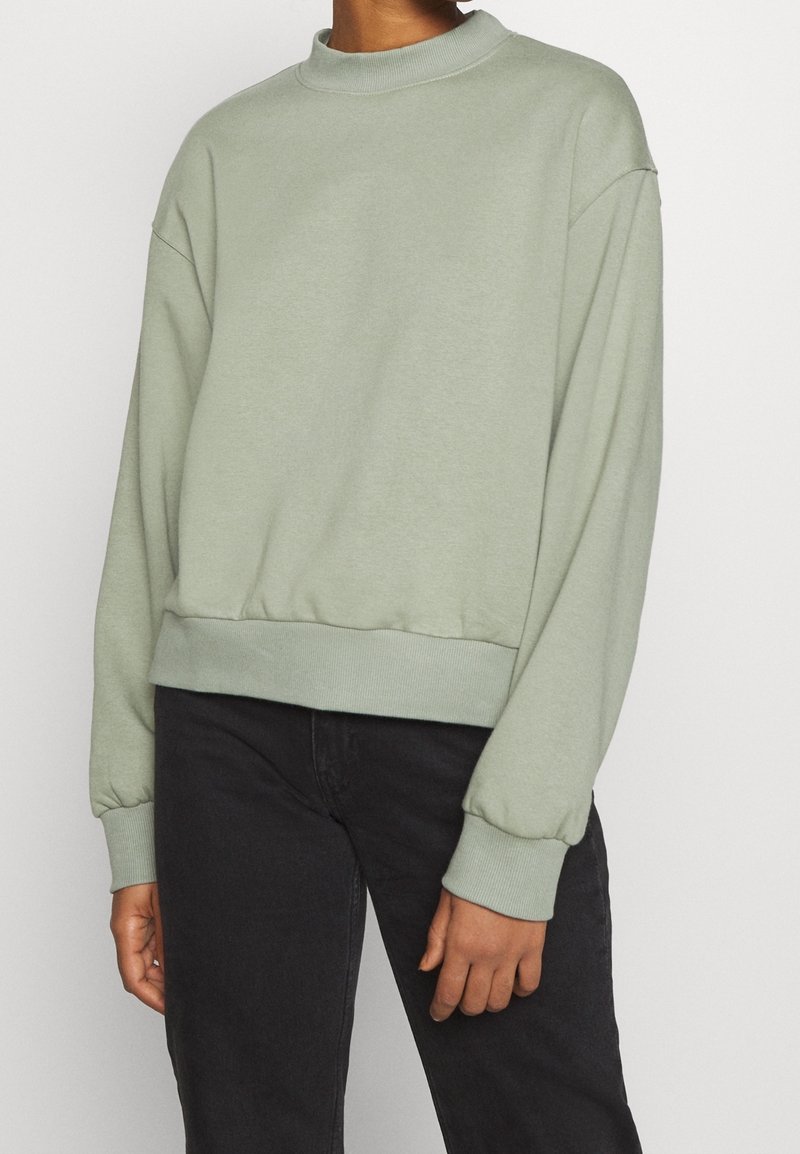 Personne portant un sweat-shirt vert clair à manches longues avec poignets et ourlet côtelés, associé à un pantalon noir sur un fond uni.