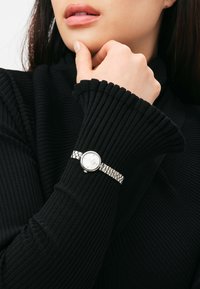 Silberne Armbanduhr mit einem runden weißen Zifferblatt, ausgestattet mit einem Metallgliederarmband, getragen auf einem schwarzen gerippten Stoffärmel. Nahaufnahme.