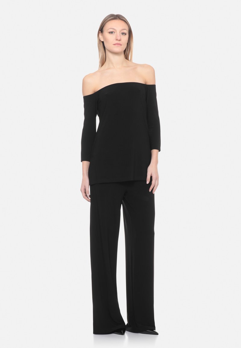 Vrouw staand, gekleed in een zwarte top zonder schouders en wijde zwarte broek, tegen een effen witte achtergrond.