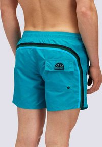 Pantaloni da bagno turchesi con bande laterali nere e blu, dotati di tasca posteriore e patch del marchio. Il materiale sembra leggero e resistente all'acqua.