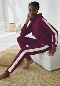 Bordeaux trainingspak met een hoodie en bijpassende broek met crème strepen langs de zijkanten. Gladde stof en een ontspannen pasvorm.