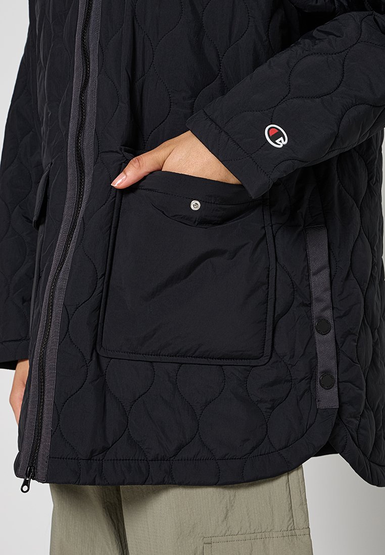 Hand in der Tasche einer schwarzen Steppjacke mit Druckknopfklappe und sichtbarem Reißverschluss, getragen mit hellen Hosen.