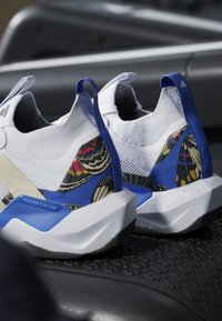 Coppia di scarpe sportive bianche e blu con dettagli a motivo di ala di farfalla sui talloni, poggiate su una superficie nera strutturata.
